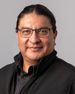 Head-and-shoulders portrait of Harley Eagle who is wearing glasses and a black zip-up jacket over a dark shirt, facing the camera with a neutral, slight smile against a plain light grey background.